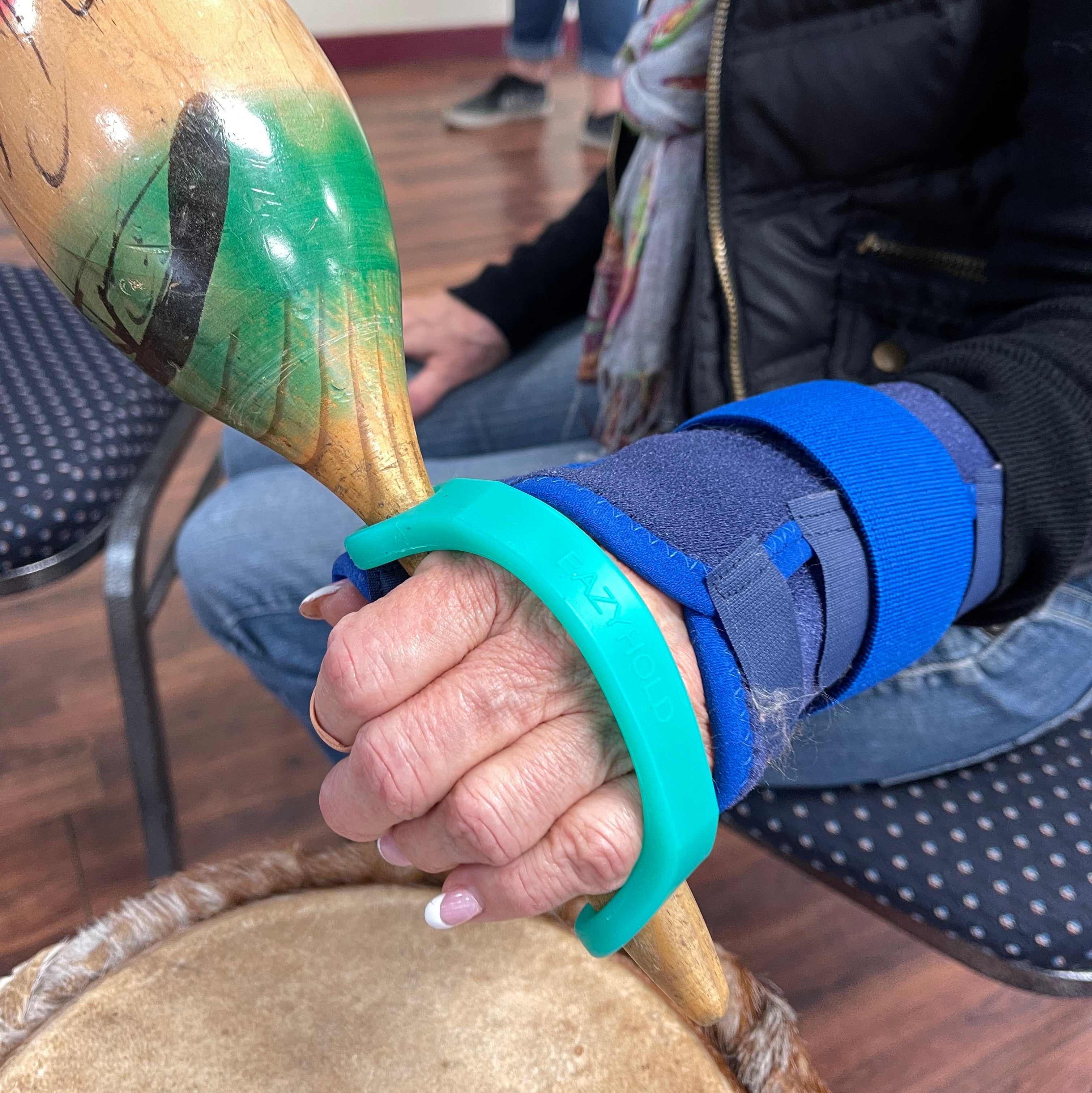 Hand with a wrist brace holding a wooden maraca with an aqua EazyHold handle.