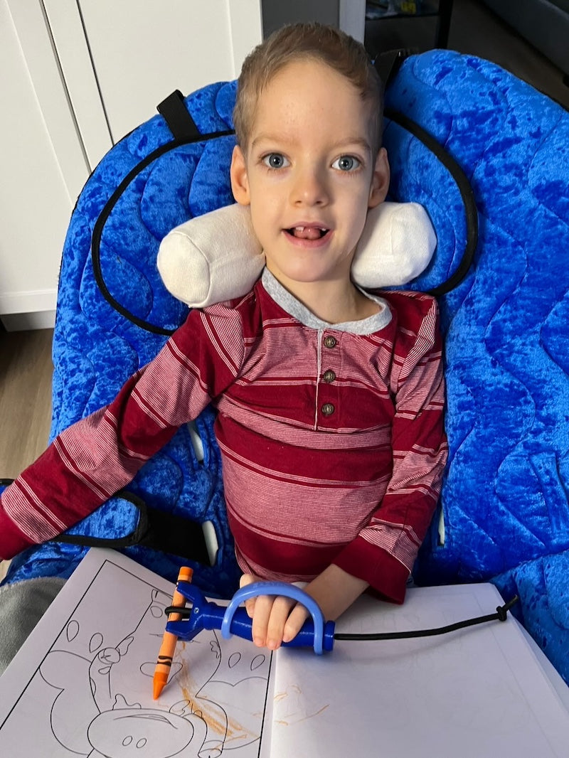 Child sitting in a blue chair with a blue EaZyHold and Functionalhand holding an orange crayon over a coloring book.