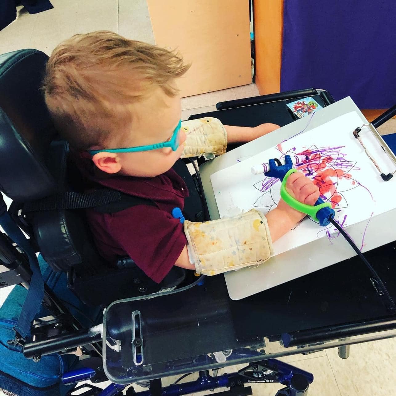 Child in a wheelchair with arm braces uses a green EaZyHold and Functionalhand to hold a marker and draw.