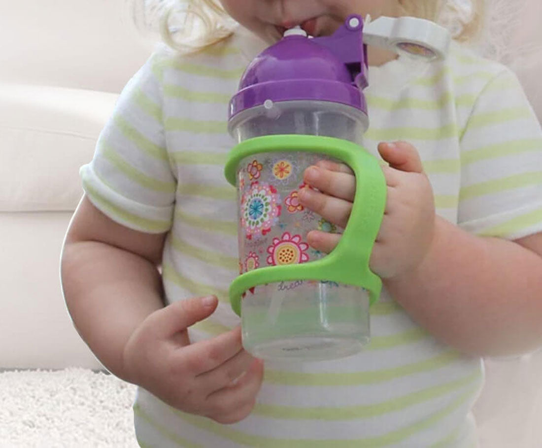 A toddler drinks from a sippy cup with a green silicone EasyHold cuff on the bottle and over her hand