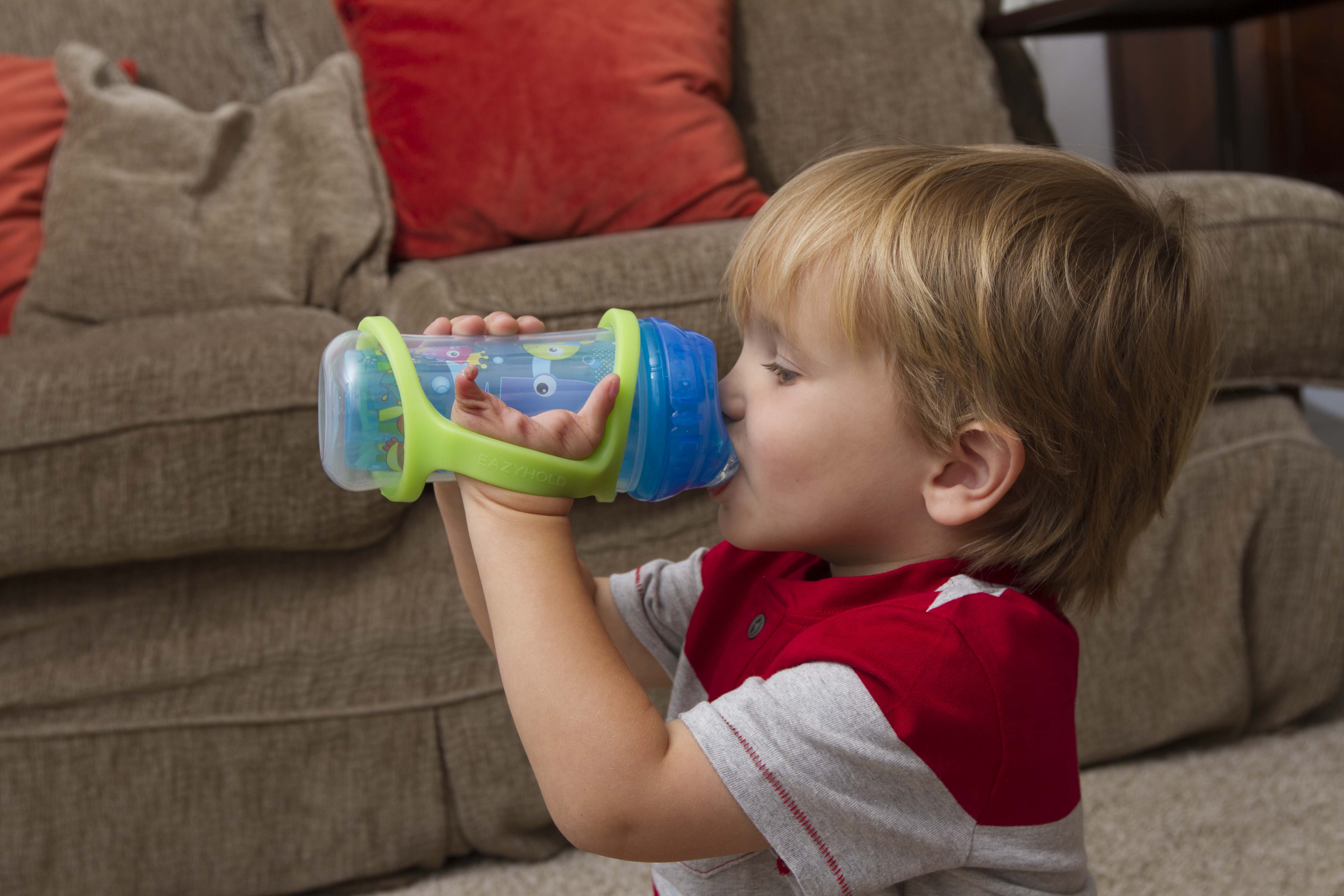 A toddler drinks from a sippy cup with a green silicone EasyHold cuff on the bottle and over his hand.