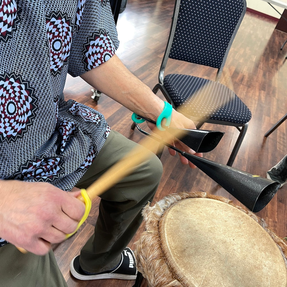 Person playing a drum with a yellow EazyHold on their drumstick and an aqua to help hold cowbells in the other hand.