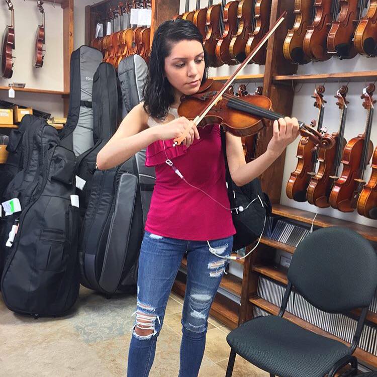 Young woman playing a violin with an orange EazyHold on her bow, in a music store with violins and cases on display.