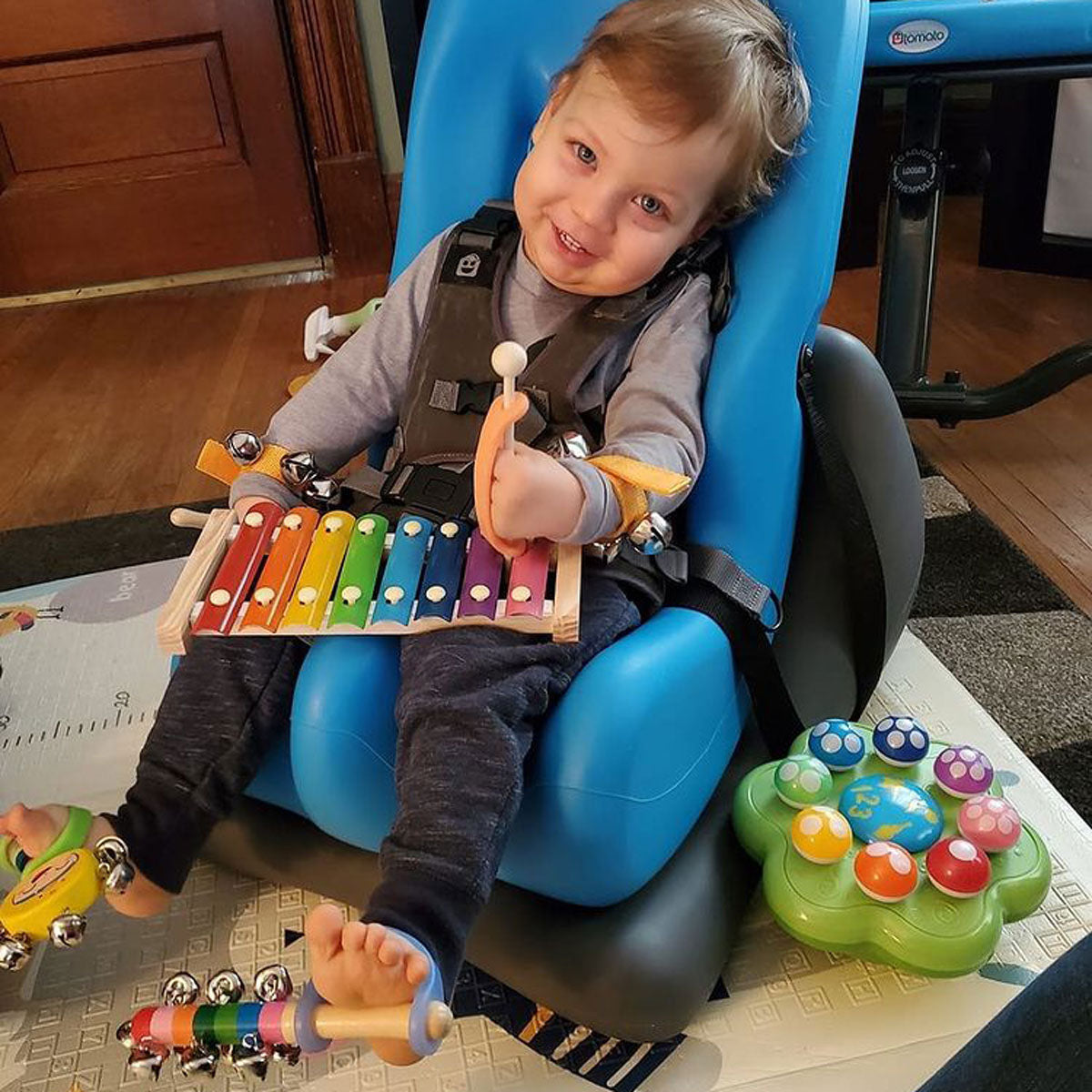 A young boy in a supportive chair with a xylophone on his lap and an orange EazyHold on his mallet. Each foot has bells with a green and blue EazyHold on them.